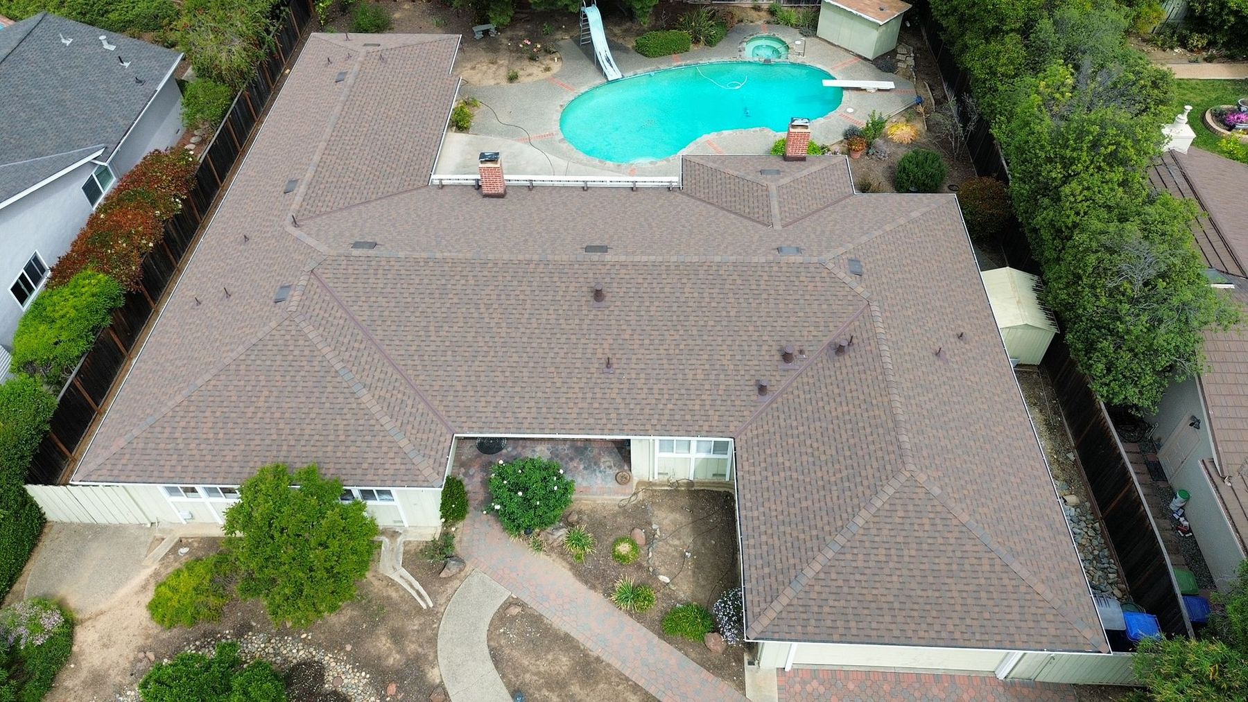 Aerial view of a completed residential shingle reroof on a Bay Area single-family home by Keith Roofing Company — warm brown architectural shingles, complex hipped roof geometry, brick chimneys, pool and patio